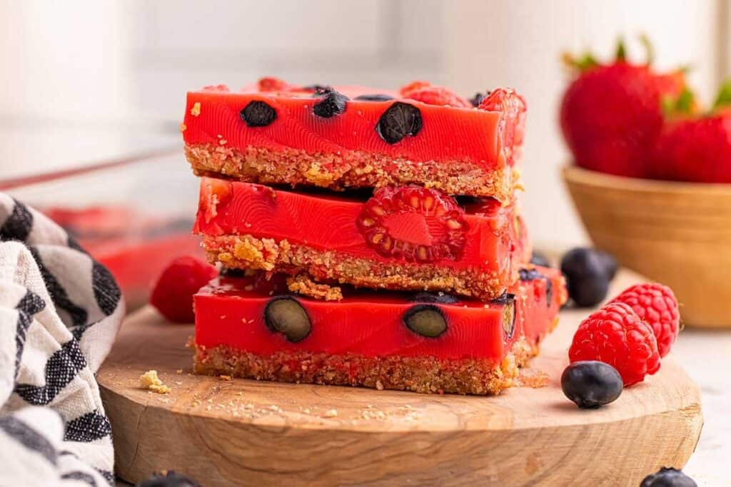 Three stacked dessert bars with a crumbly base and a red gelatin layer containing raspberries and blueberries, placed on a wooden board with loose berries around.