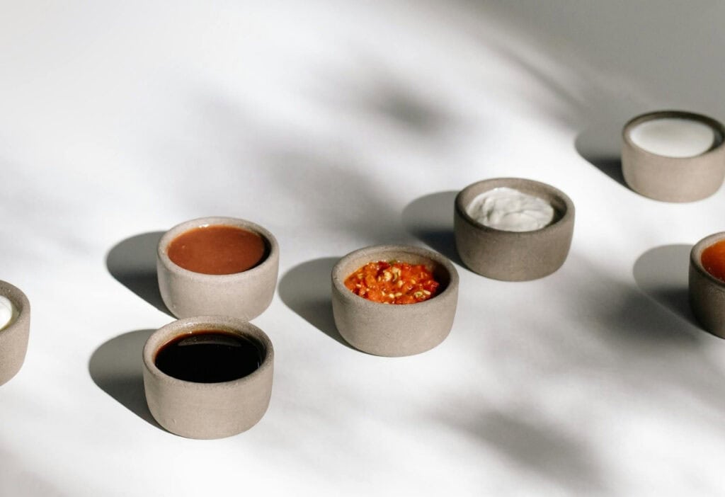 Six small ceramic bowls, each containing a different sauce or dip, arranged on a white surface with soft shadows.