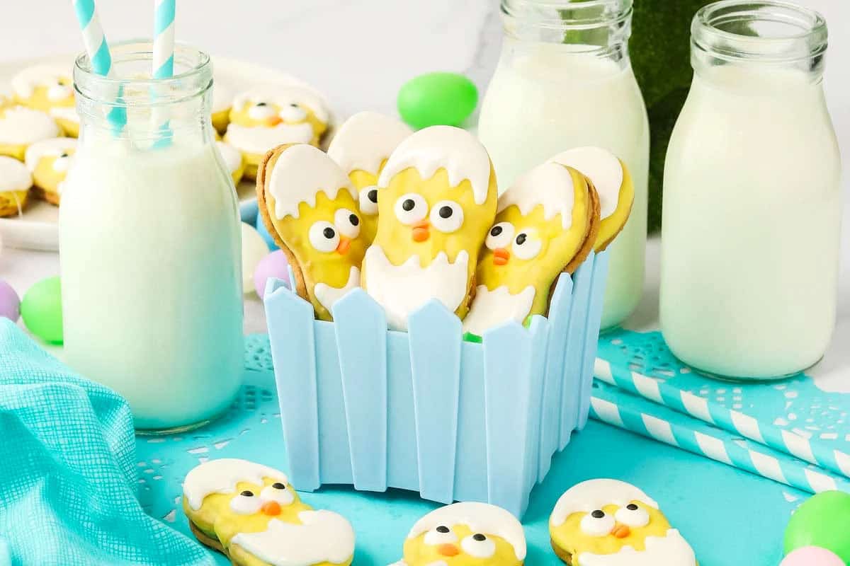 Cookies decorated as chicks hatching from eggs are arranged in a small blue fence-style container, surrounded by milk bottles, straws, and pastel eggs on a table.