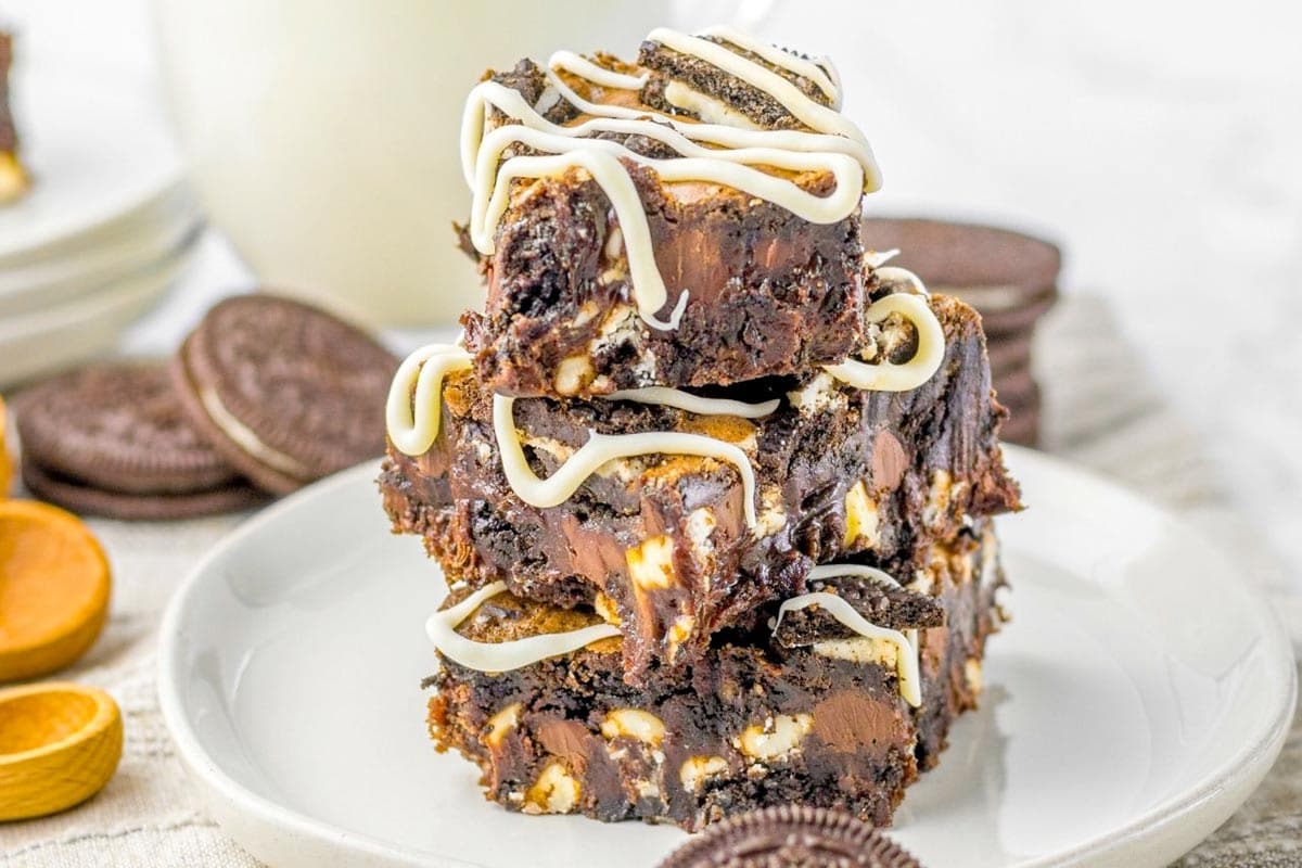 Three chocolate brownies with white chocolate drizzle are stacked on a white plate, surrounded by sandwich cookies and chocolate pieces.