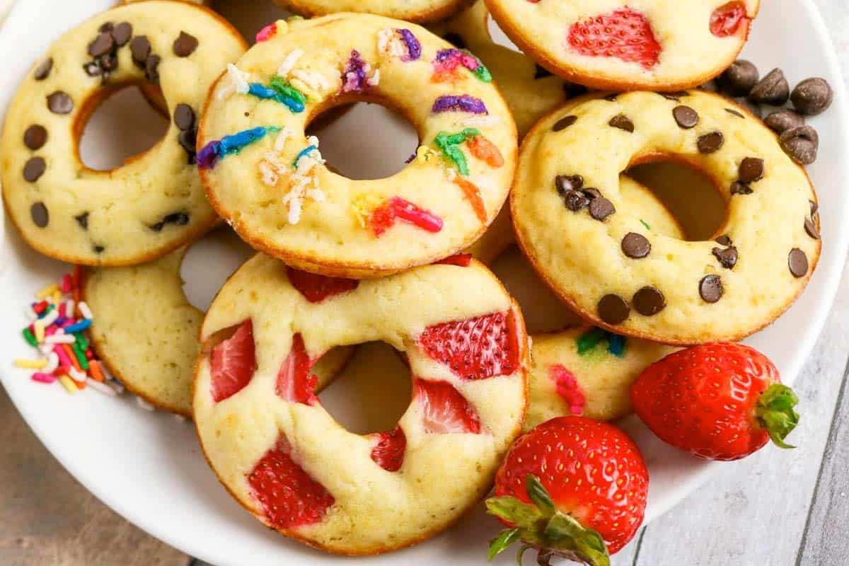 A plate of baked donut-shaped treats topped with chocolate chips, rainbow sprinkles, and pieces of strawberry, with two whole strawberries on the side.