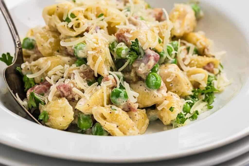 A close-up of pasta shells mixed with peas, ham, creamy sauce, and shredded cheese, garnished with parsley in a white bowl.