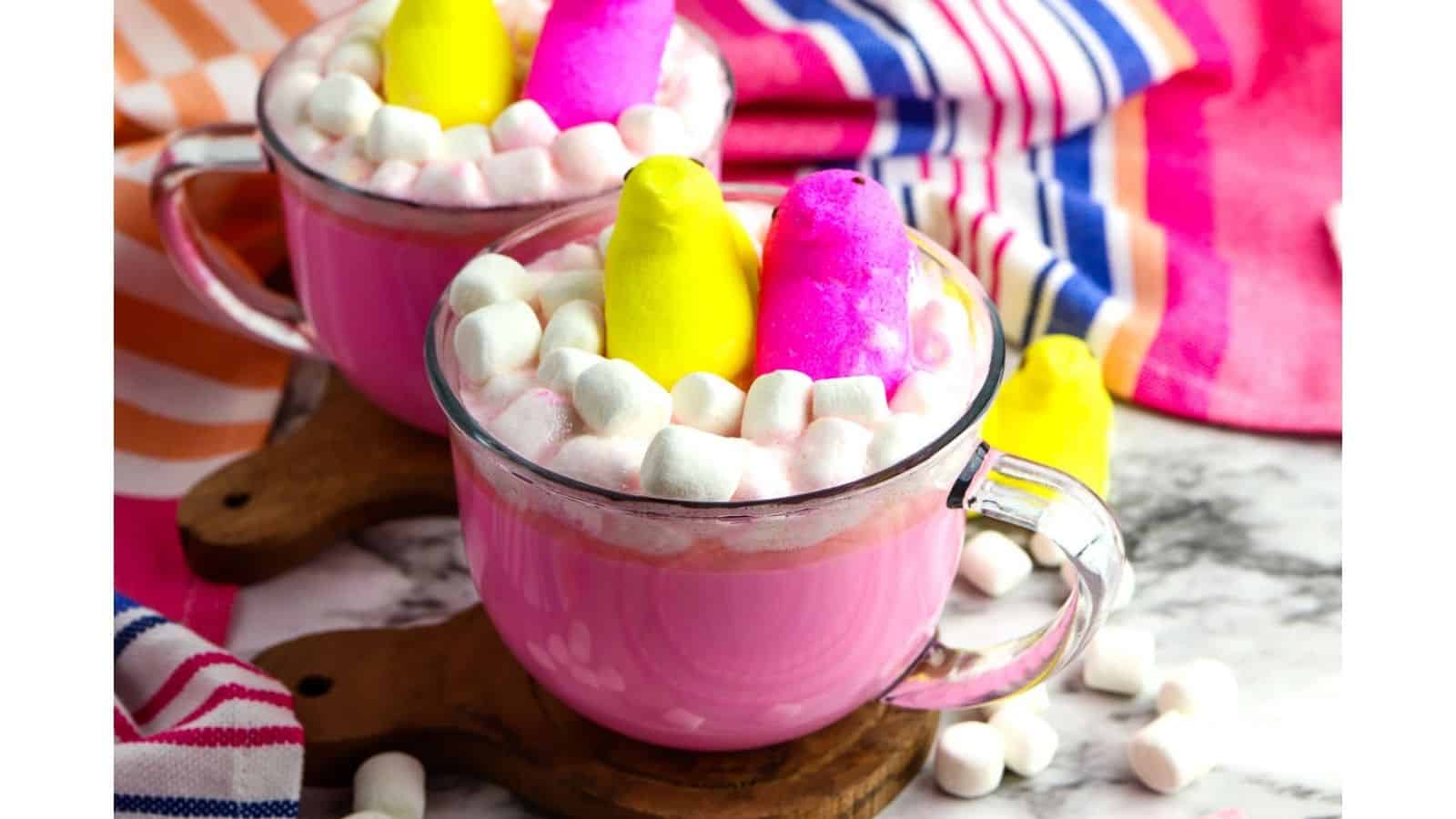 Two glass mugs of pink hot chocolate topped with pastel-colored marshmallow chicks and mini marshmallows, set on wooden coasters. Striped fabric in the background.
