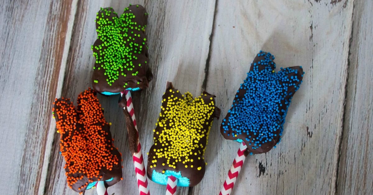 Four marshmallow bunnies on sticks, coated in chocolate and topped with orange, green, yellow, and blue sprinkles, are arranged on a wooden surface.