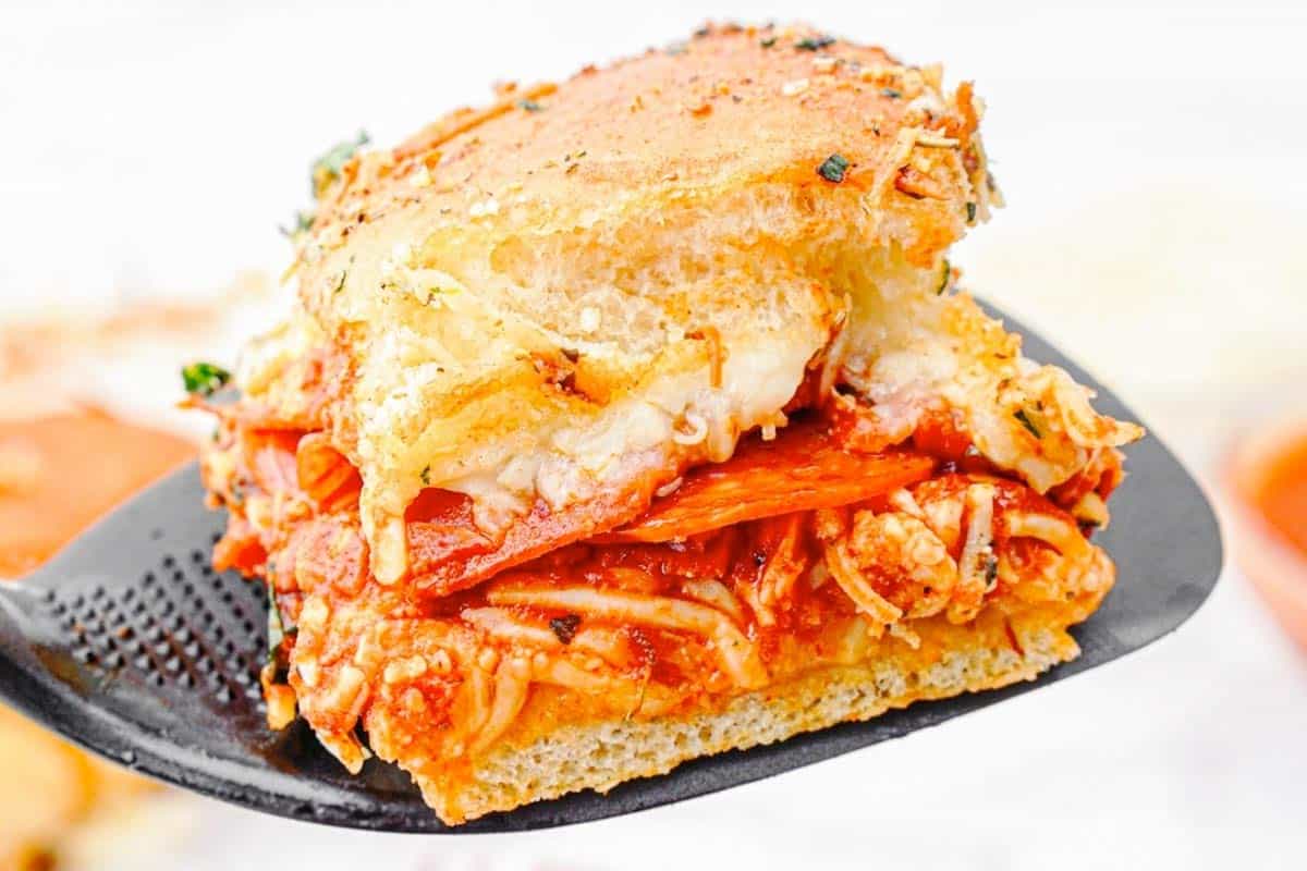 A close-up of a spatula holding a square piece of baked dish with melted cheese, tomato sauce, pepperoni, and herbs on golden bread, with more pieces visible in the blurred background.