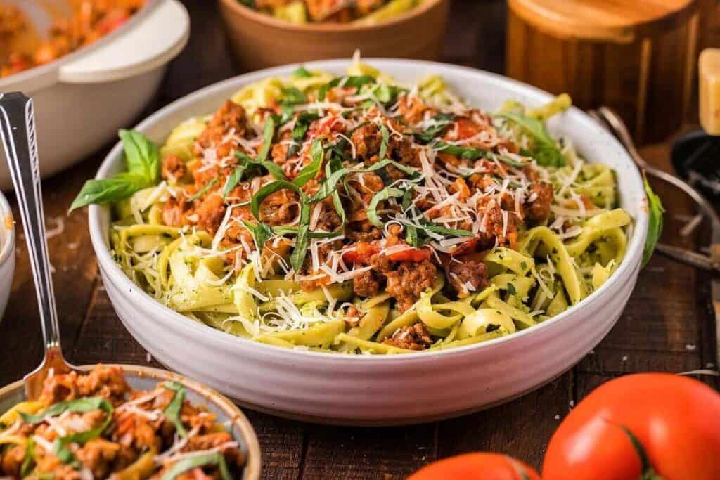 A bowl of fettuccine pasta topped with meat sauce, fresh basil, and grated cheese sits on a wooden table alongside tomatoes and other dishes.