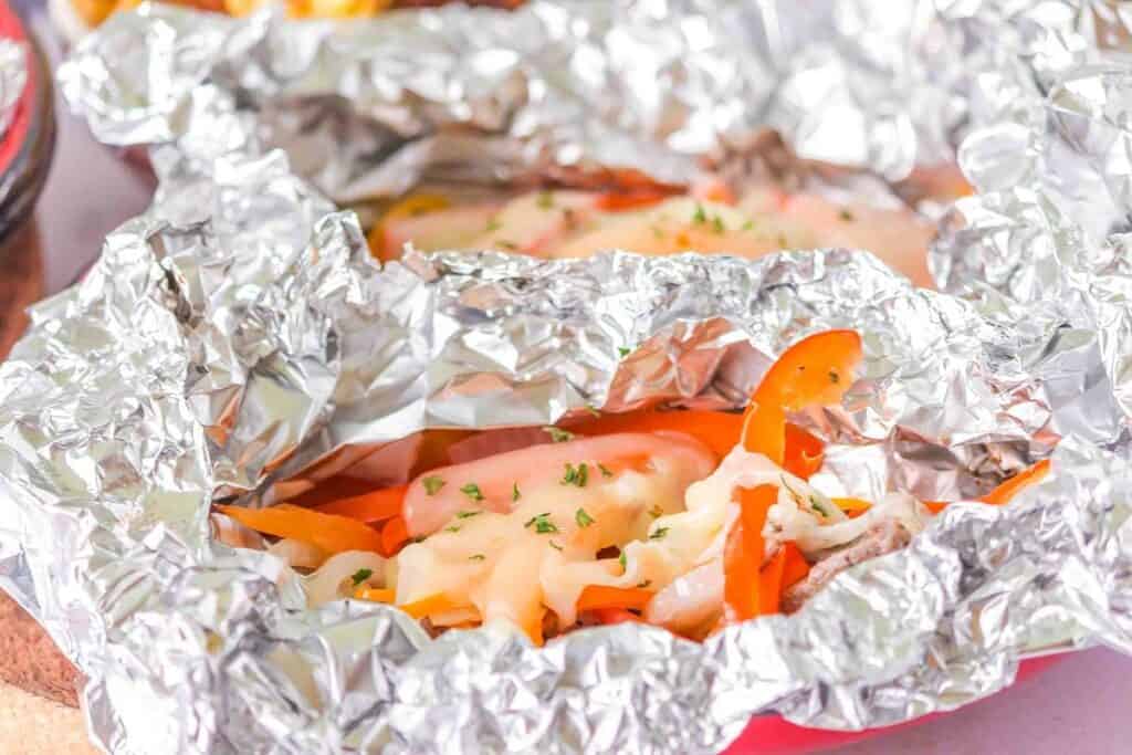 Close-up of cooked vegetables and melted cheese in an open aluminum foil packet, garnished with chopped herbs.