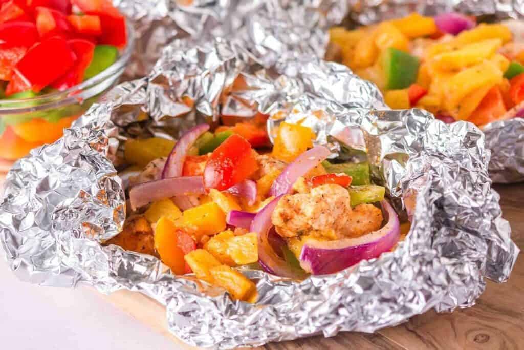 Colorful chopped vegetables and seasoned chicken are cooked together in an open foil packet, with a bowl of additional chopped vegetables in the background.
