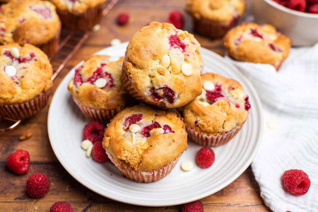 A plate of raspberry and white chocolate muffins sits on a wooden table, surrounded by fresh raspberries and a wire cooling rack.