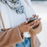 A person wearing a brown fleece jacket and a scarf is using a smartphone outdoors. Only their hands and torso are visible in the image.