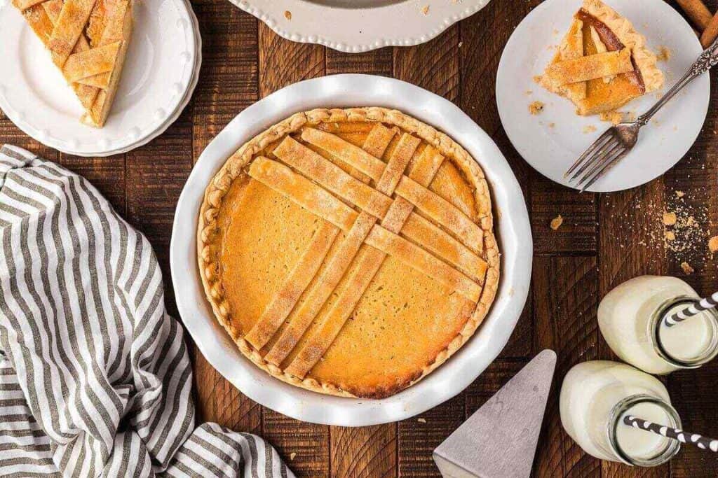 Top-down view of a baked pie with a lattice crust in a white dish, with slices served on plates, striped napkin, milk bottles, and utensils on a wooden table.