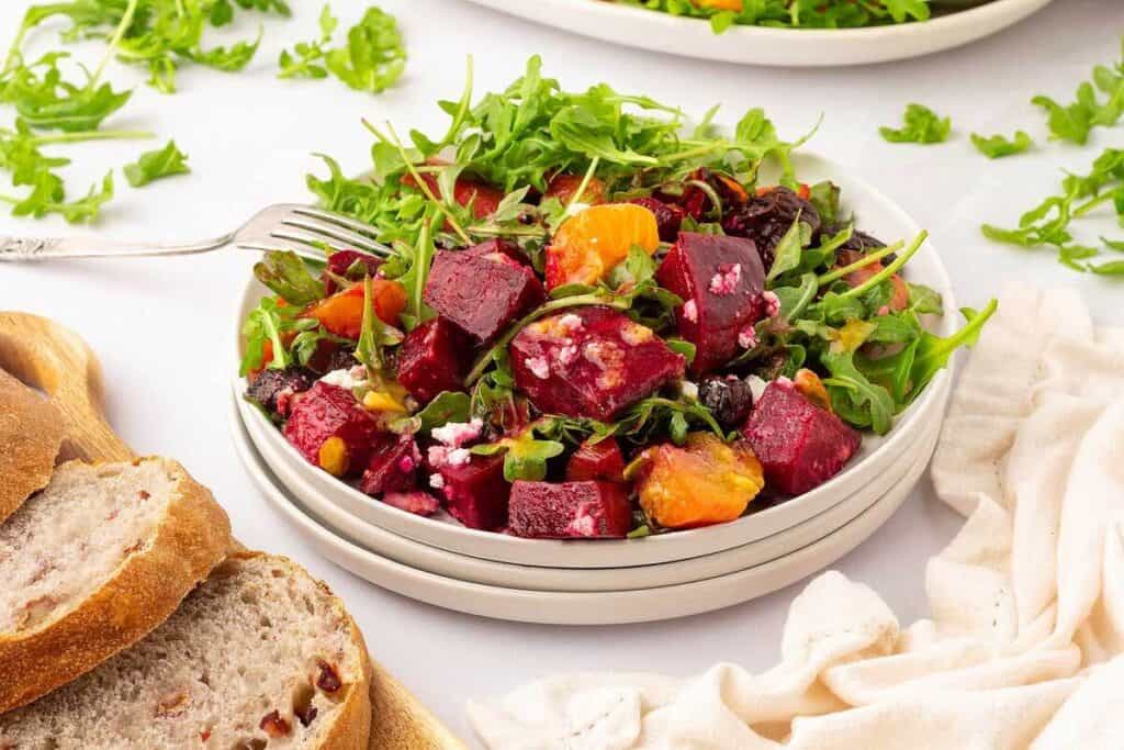 A plate of salad with arugula, roasted beets, orange segments, nuts, and cheese, served alongside slices of bread on a wooden board.