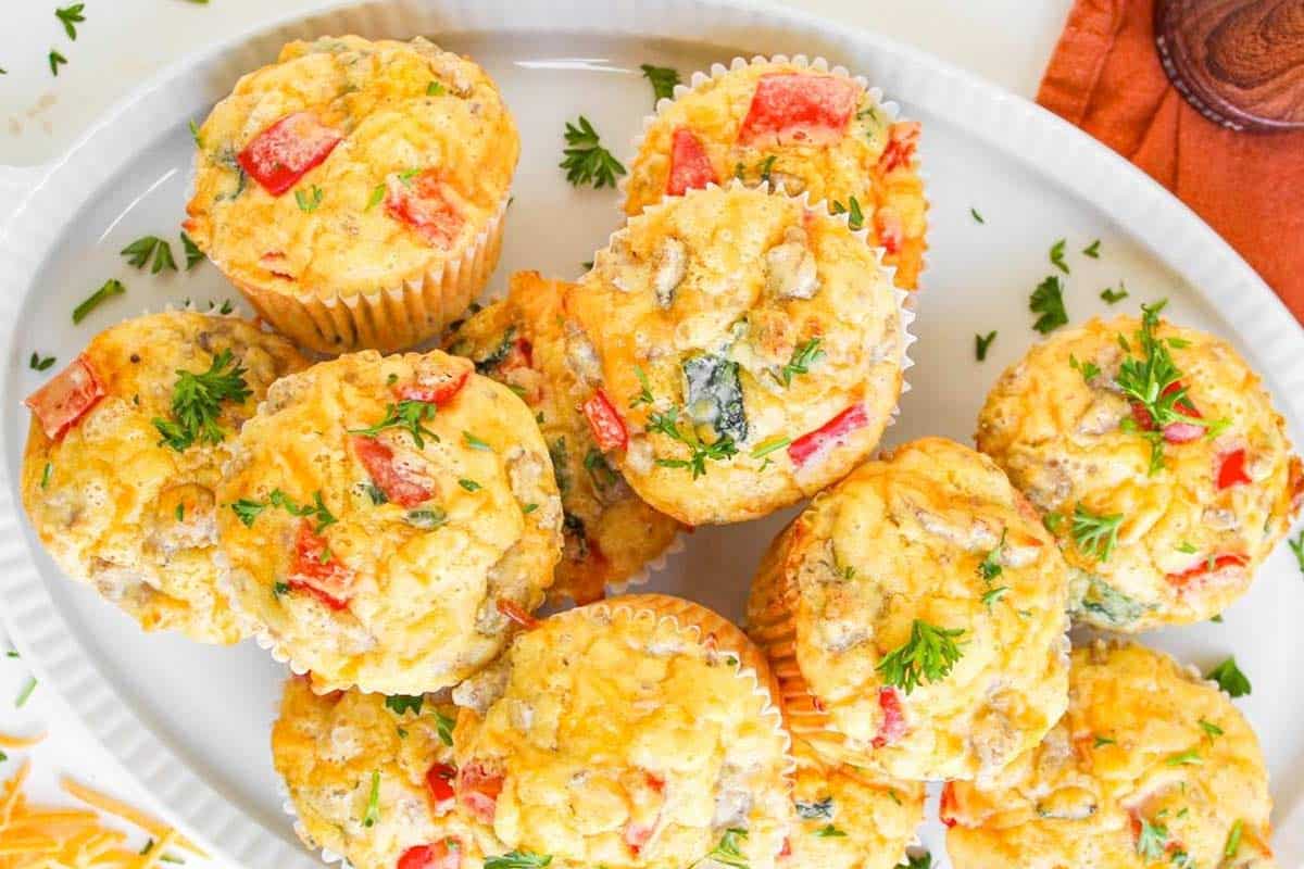 A white platter with several savory muffins containing visible pieces of red bell pepper, green herbs, and possibly cheese, garnished with fresh herbs.