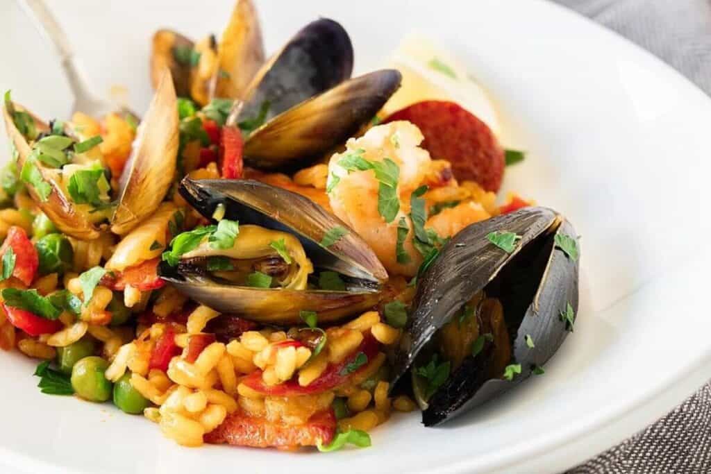 A plate of seafood paella with mussels, shrimp, rice, peas, red peppers, and garnished with chopped parsley.