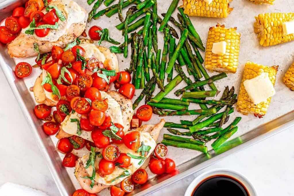 Sheet pan with baked chicken breasts topped with tomatoes and basil, roasted asparagus, and corn on the cob with butter, next to a small bowl of dark sauce.