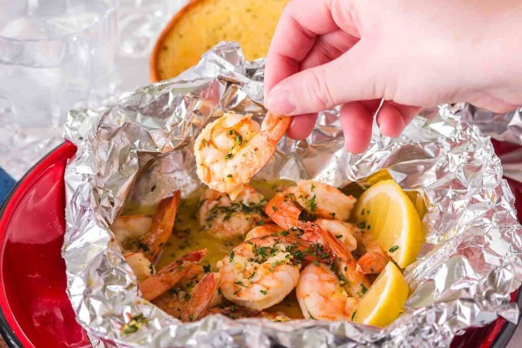A hand picks up a cooked shrimp from a foil packet filled with shrimp, herbs, and lemon wedges, with a slice of bread in the background.