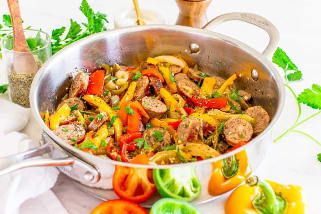 Stainless steel pan filled with cooked sausage slices, red and yellow bell peppers, onions, and herbs, surrounded by fresh vegetables and seasonings on a white surface.