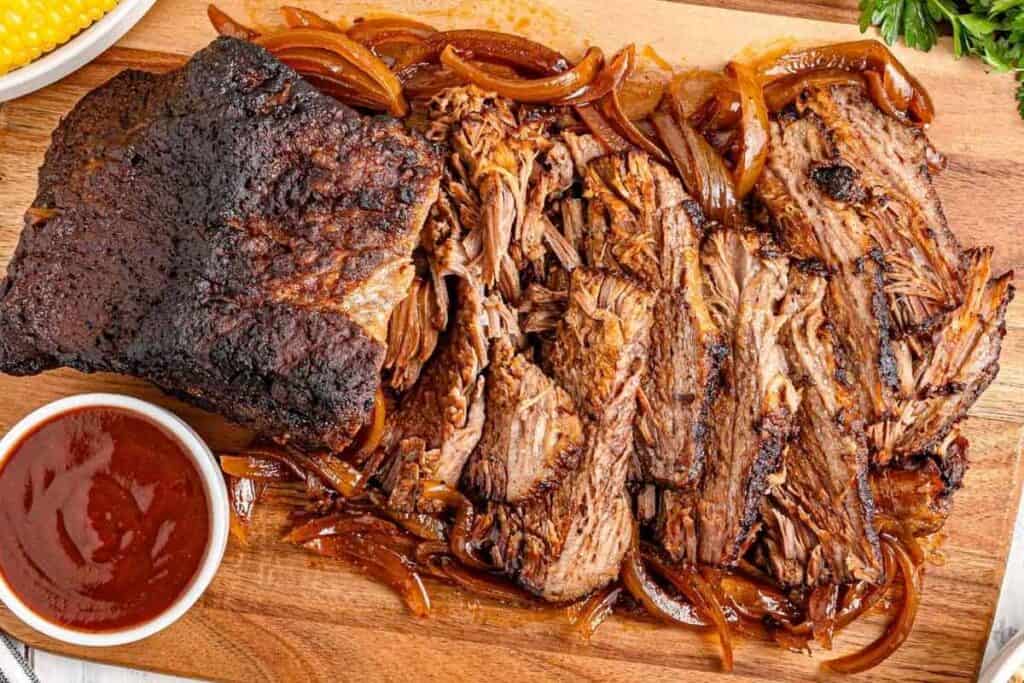 Sliced and shredded beef brisket with caramelized onions on a wooden cutting board, served with a cup of barbecue sauce.