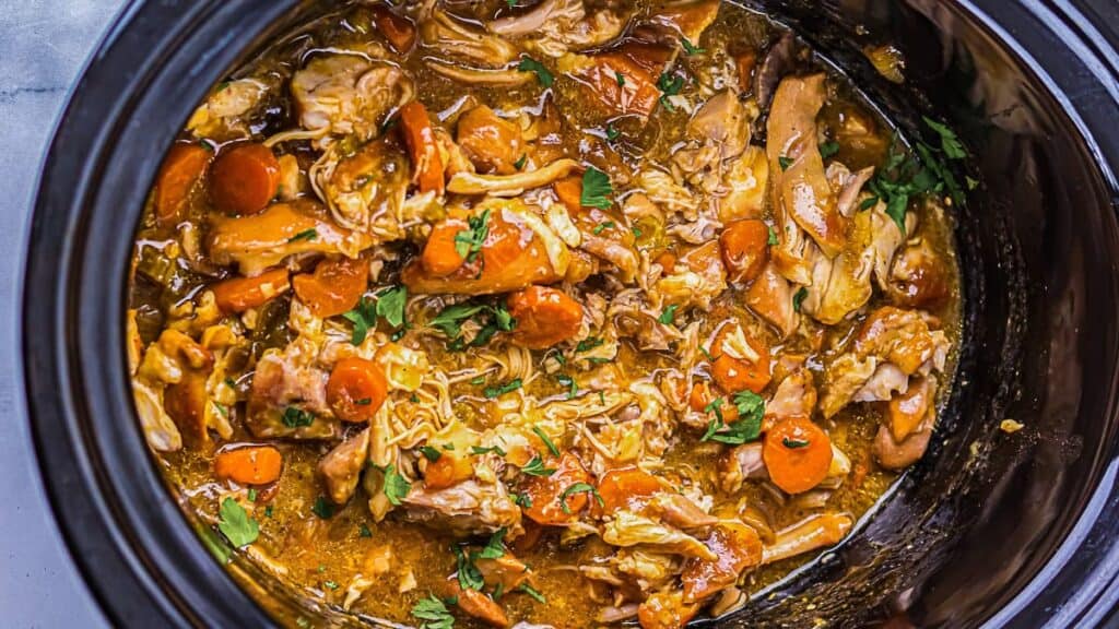 Close-up of a slow cooker filled with shredded chicken, sliced carrots, and chopped herbs in a thick, saucy broth.
