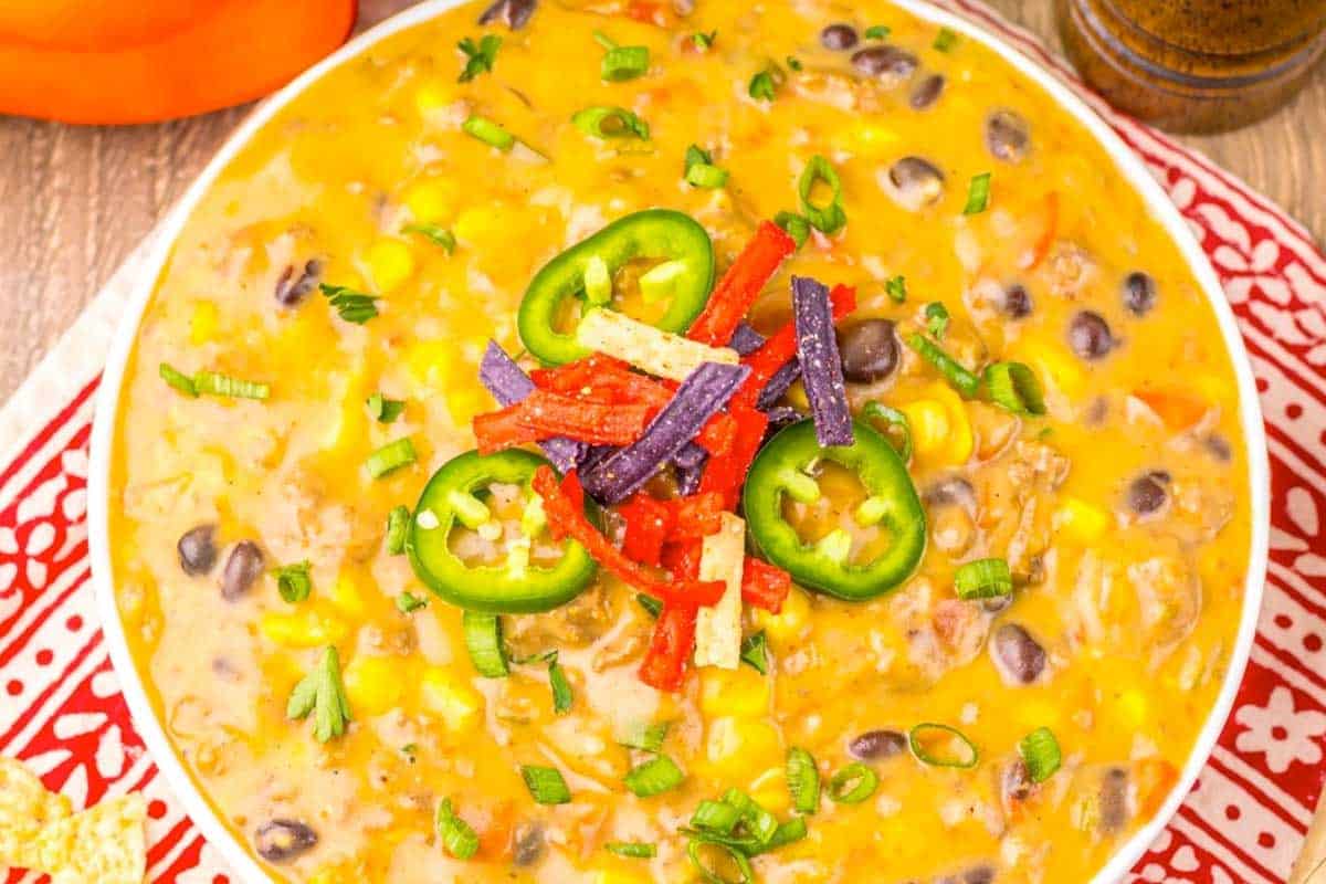 A bowl of creamy soup with black beans, corn, and ground meat, topped with sliced jalapeños, tortilla strips, and chopped green onions, on a red patterned cloth.