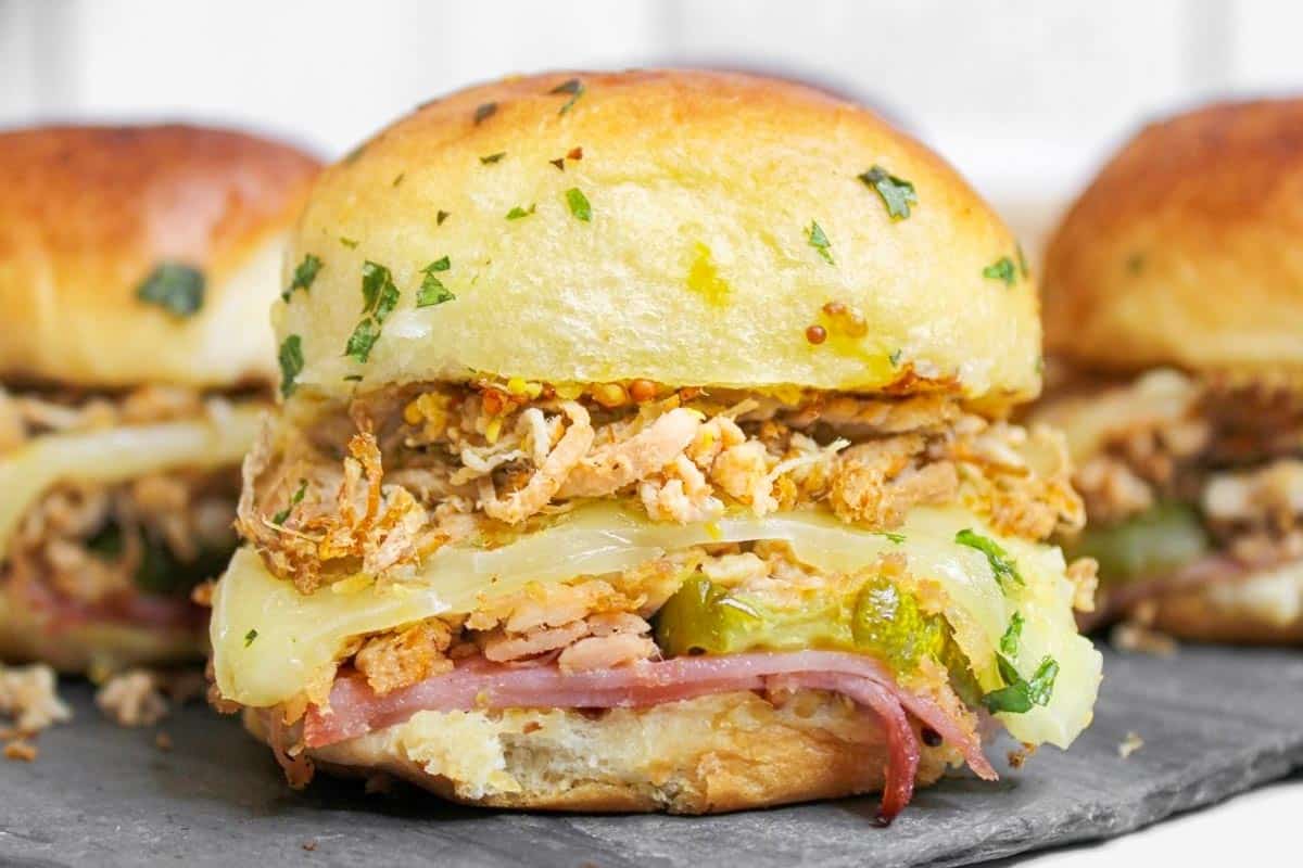 Cuban slider sitting on a black cutting. board with more Cuban sliders in the background.