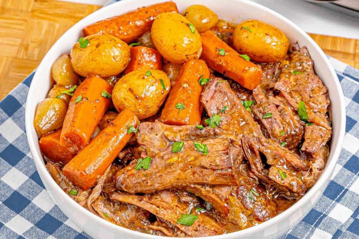 A white plate holds Crockpot Pot Roast, shredded and surrounded by whole baby potatoes and cooked carrots in a brown sauce, garnished with chopped parsley. The plate is set on woven placemats with a striped napkin nearby.