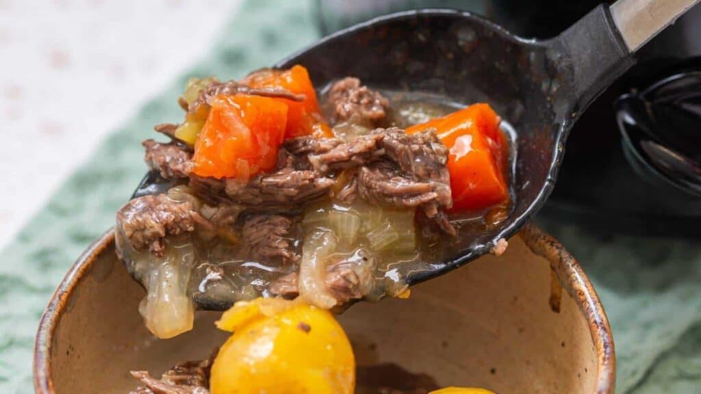 A ladle serving beef stew with chunks of beef, carrots, onions, and potatoes into a bowl.