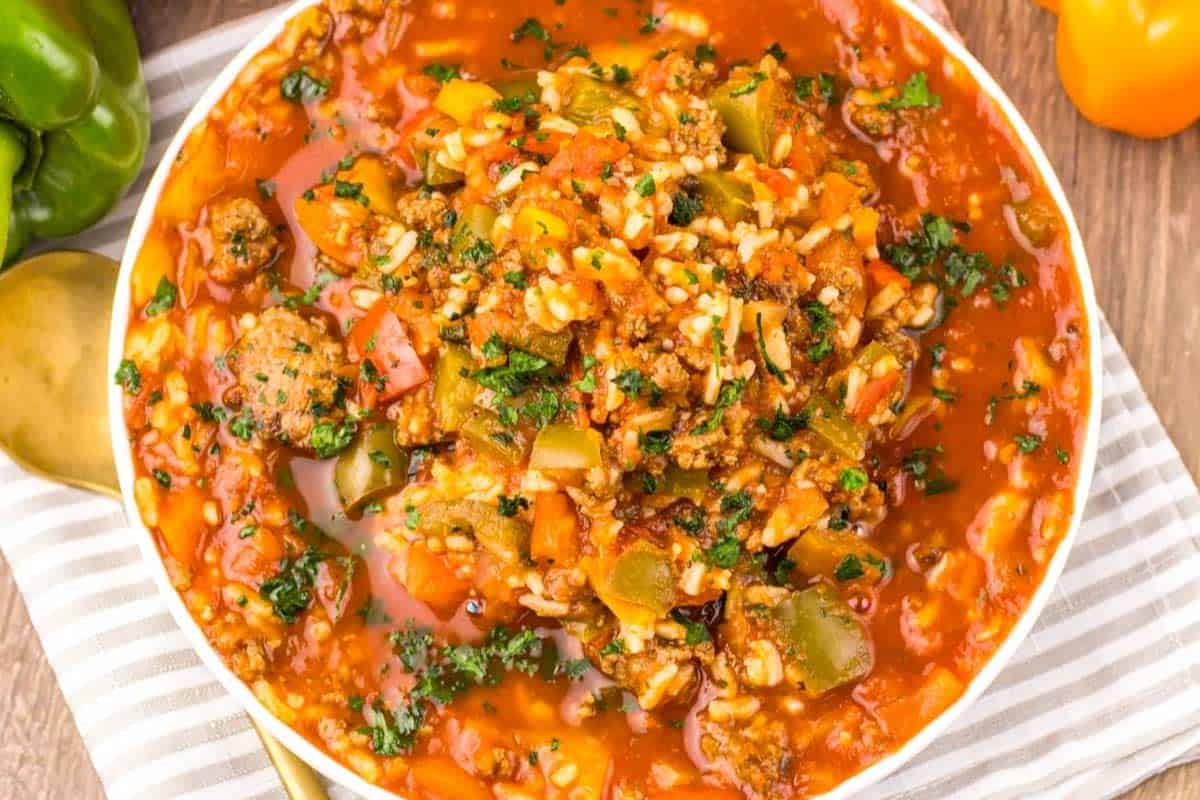 A bowl of Crockpot Stuffed Pepper Soup with ground meat, rice, green and red bell peppers, and a tomato-based broth, garnished with herbs, sits on a striped cloth next to fresh green and yellow bell peppers.
