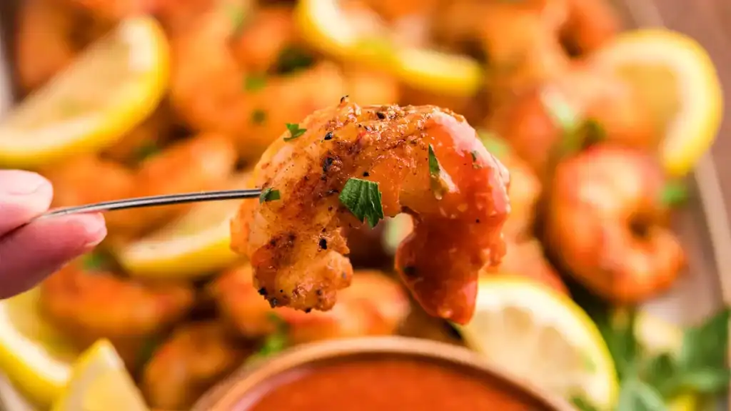 A hand holds a fork with a piece of grilled shrimp garnished with herbs, with a bowl of cocktail sauce and lemon slices in the background.