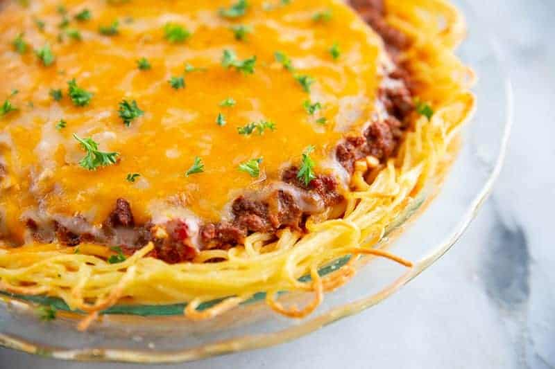 A baked spaghetti pie with a cheesy top layer, meat sauce, and garnished with chopped parsley in a glass pie dish.