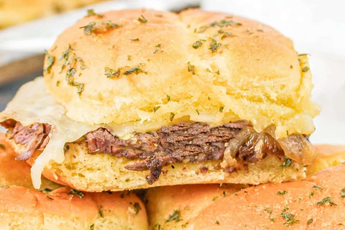 A close-up of a steak slider with a toasted bun, filled with sliced beef, melted cheese, and caramelized onions. The bun is sprinkled with herbs and the sandwich rests on a tray with other tempting sliders in the background.