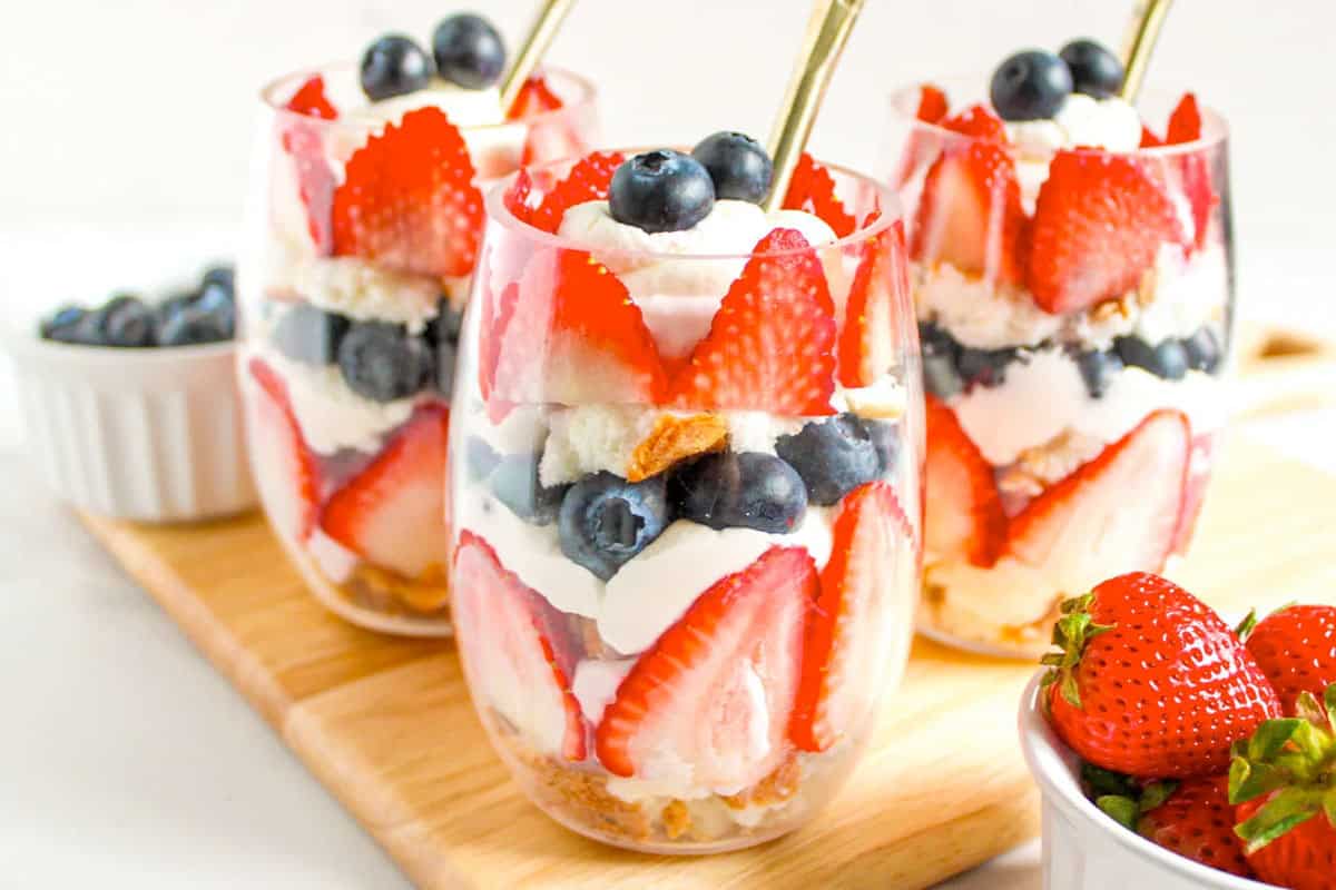 Three clear glasses filled with layered trifles of strawberries, blueberries, whipped cream, and angel food cake, with spoons in each glass. Bowls of strawberries and blueberries are nearby.