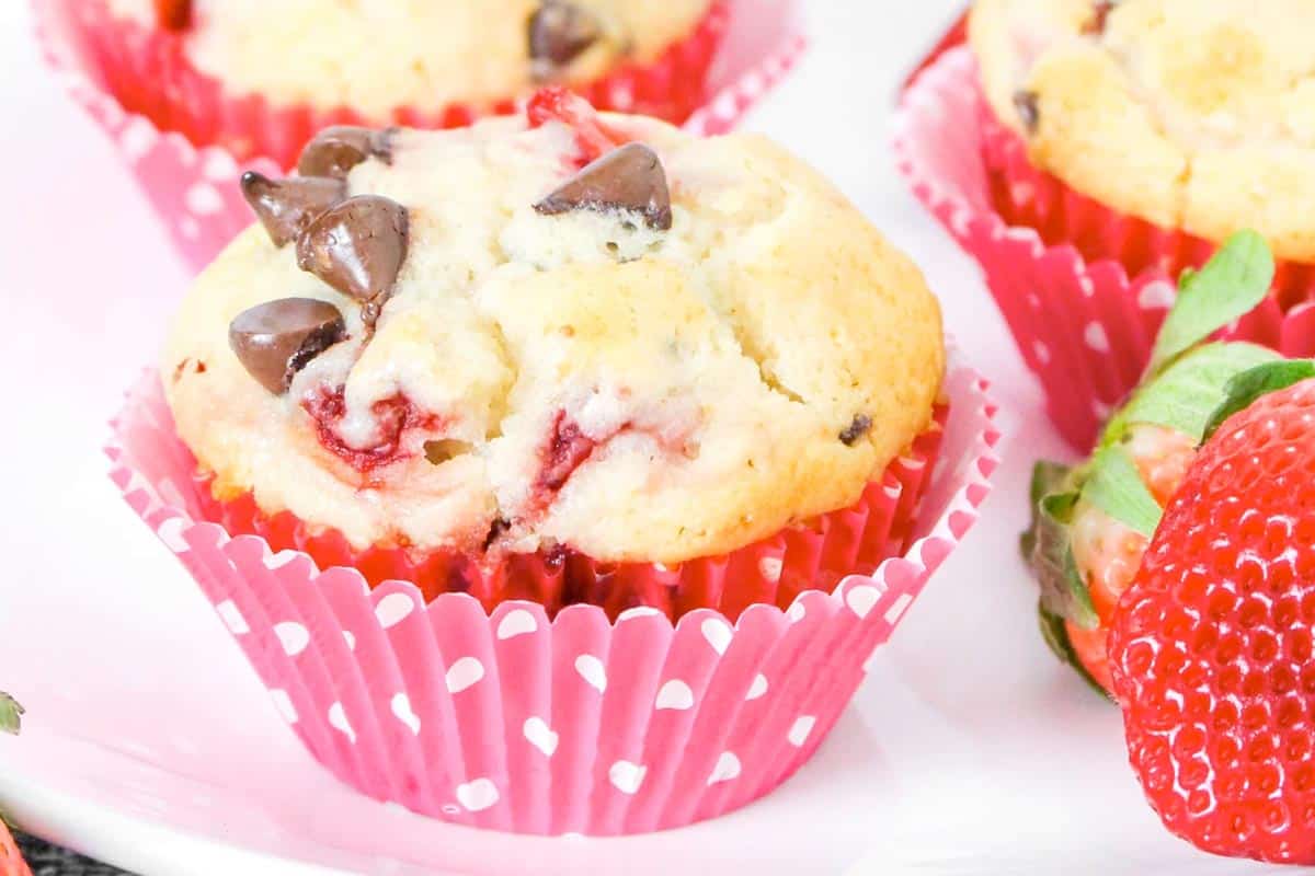 A strawberry chocolate chip muffin in a pink polka dot wrapper, with a fresh strawberry beside it on a white surface.