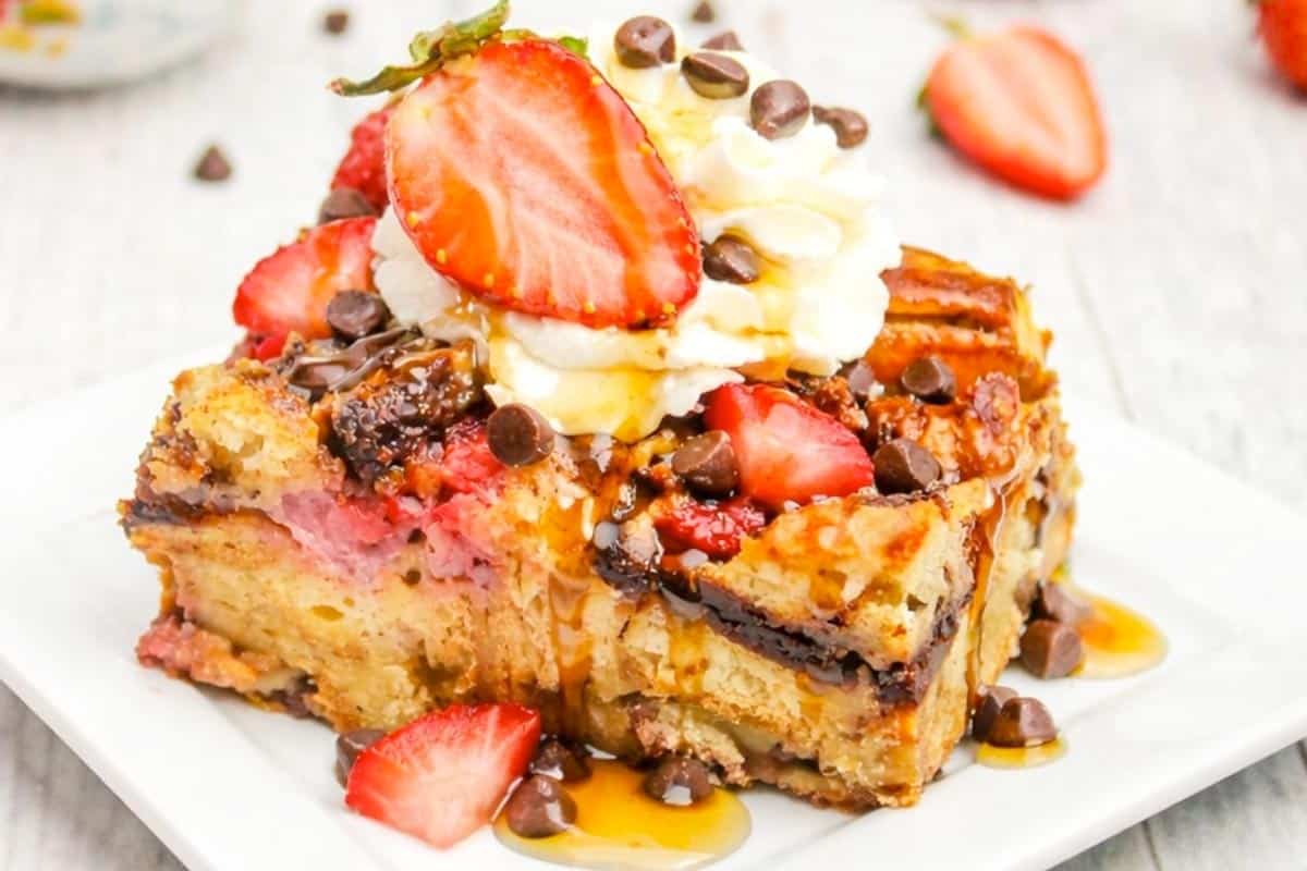 A slice of baked French toast topped with whipped cream, sliced strawberries, chocolate chips, and a drizzle of syrup on a white plate.
