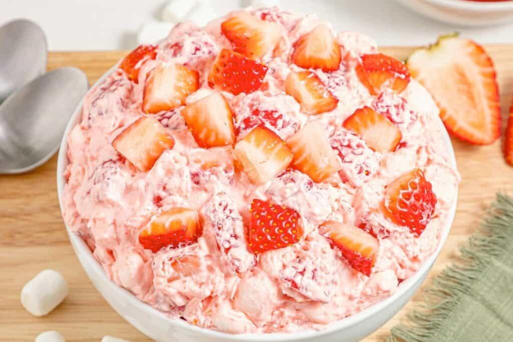 A bowl of strawberry fluff salad with chopped strawberries on top, placed on a wooden surface next to two spoons, marshmallows, and a green napkin.