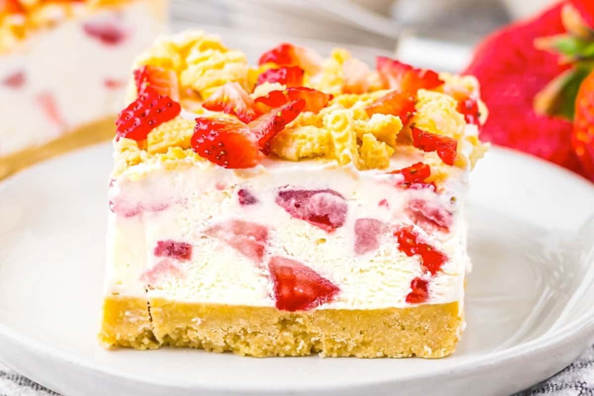 A square slice of strawberry shortcake ice cream bar with visible strawberry pieces and a crumbly topping, served on a white plate.