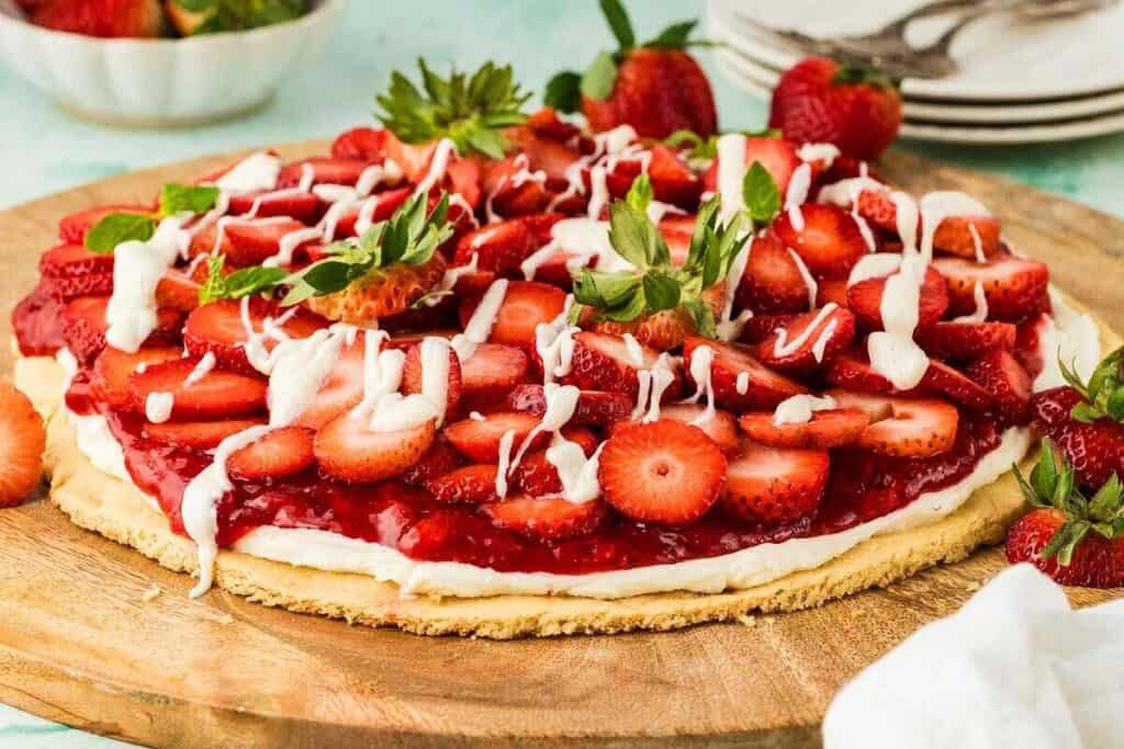 A large cookie crust topped with cream, strawberry glaze, fresh sliced strawberries, and drizzled white icing sits on a wooden board.