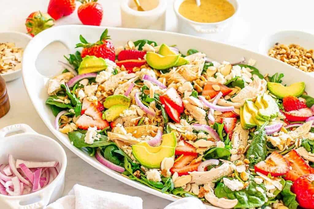 A large white dish contains a salad with spinach, sliced strawberries, avocado, red onion, chicken, nuts, and crumbled cheese. Surrounding bowls hold more salad ingredients and dressing.