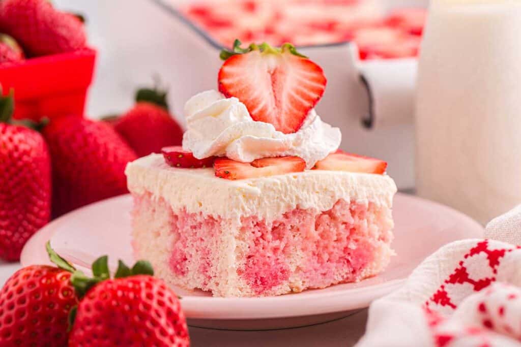 A slice of strawberry poke cake topped with whipped cream and fresh strawberry slices on a pink plate, with whole strawberries in the background.