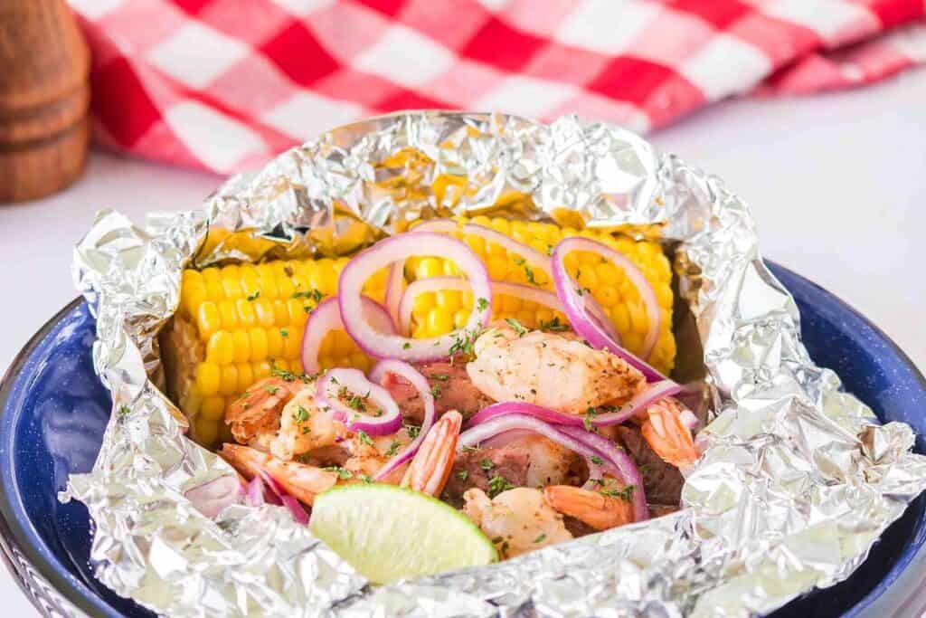 Shrimp, corn on the cob, and sliced red onions served in a foil packet with a lime wedge, on a blue plate, with a red and white checkered cloth in the background.