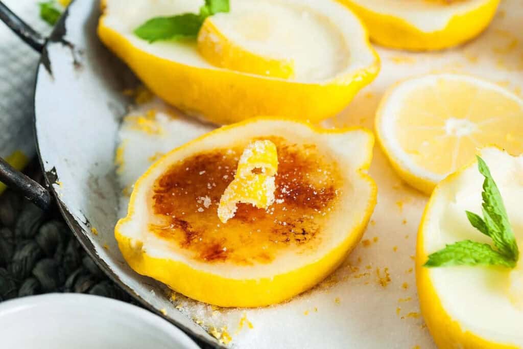 Halved lemons filled with custard, topped with caramelized sugar and garnished with sugared lemon peel and mint leaves on a white plate&mdash;a refreshing twist on classic citrus desserts.