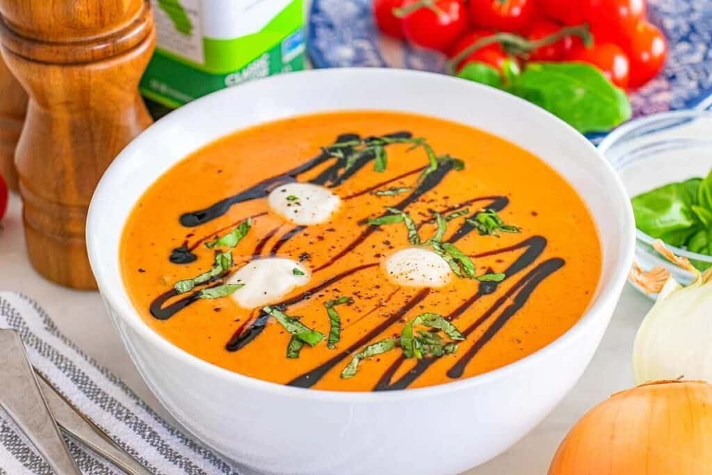 A bowl of creamy tomato soup topped with cream, chopped basil, and a drizzle of balsamic glaze, surrounded by fresh vegetables and soup ingredients.
