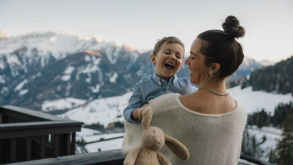 A woman holds a smiling young child and a stuffed bunny on a snowy mountain balcony, with snow-covered peaks in the background.