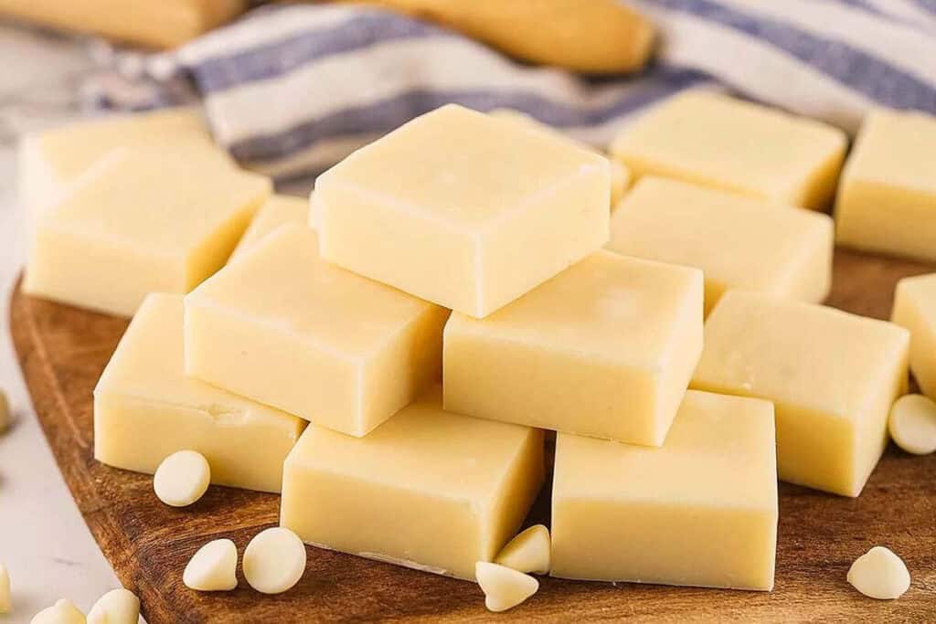 Squares of white chocolate fudge are stacked on a wooden board, with scattered white chocolate chips and a striped cloth in the background.