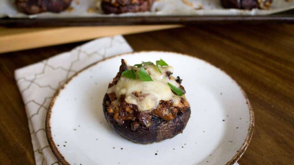 A stuffed mushroom topped with melted cheese and herbs is served on a white plate, with more stuffed mushrooms visible on a tray in the background.