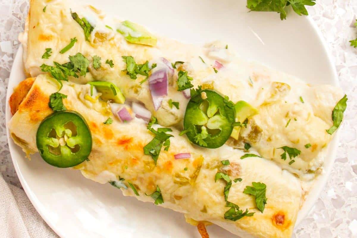 Two enchiladas topped with melted cheese, sliced jalape&ntilde;os, chopped red onion, and fresh cilantro sit on a white plate. The surface underneath is a speckled, light-colored countertop.