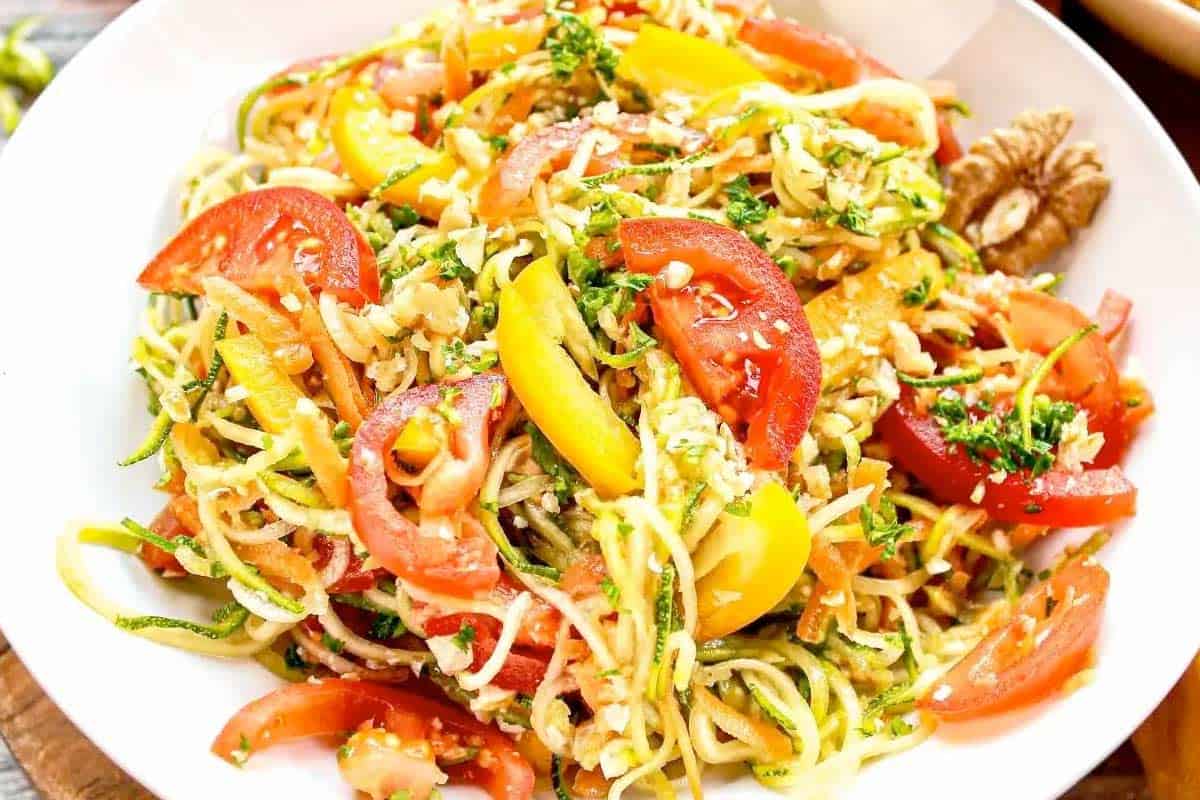 A bowl of zucchini noodles mixed with cherry tomatoes and yellow bell peppers makes a refreshing zucchini salad, garnished with herbs, with fresh veggies and a fork nearby.