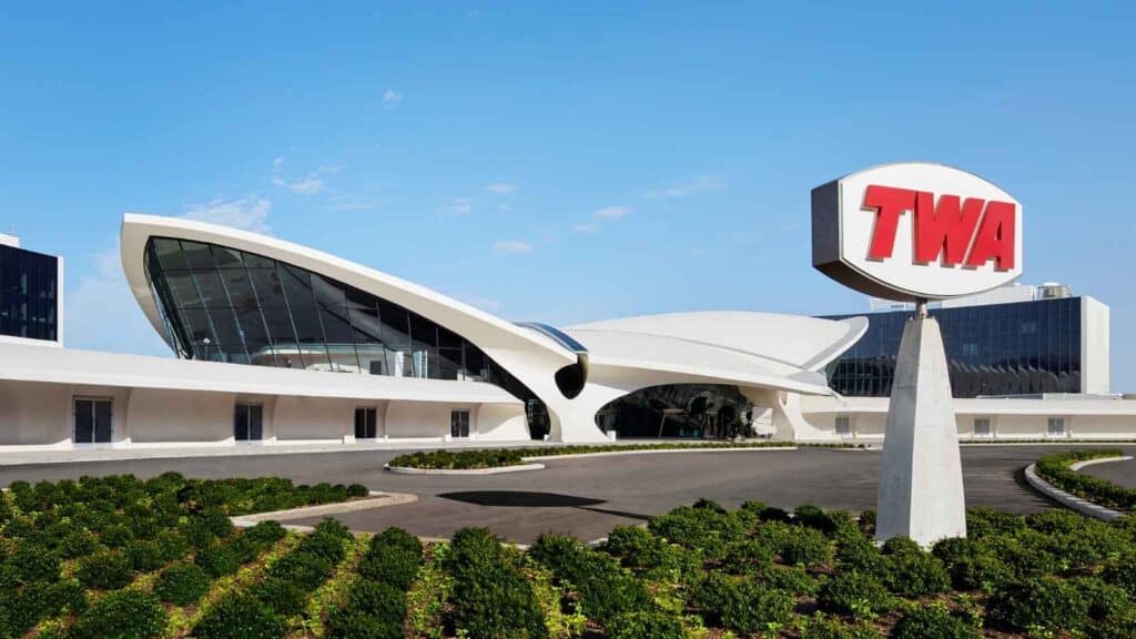 The TWA Hotel entrance at JFK Airport features retro architecture, large glass windows, and a vintage TWA sign in front of the building.