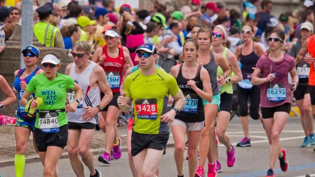 A group of runners wearing numbered bibs participates in a road race reminiscent of the Boston Marathon, with spectators and officials visible in the background.