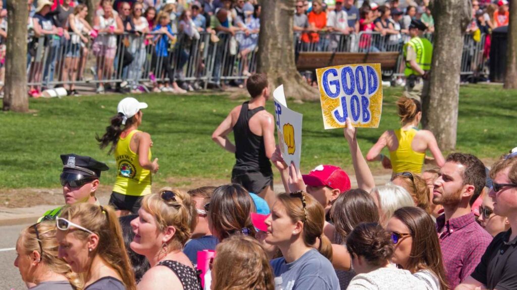 A crowd of spectators at a marathon cheers on runners; one person holds a sign that reads "GOOD JOB.